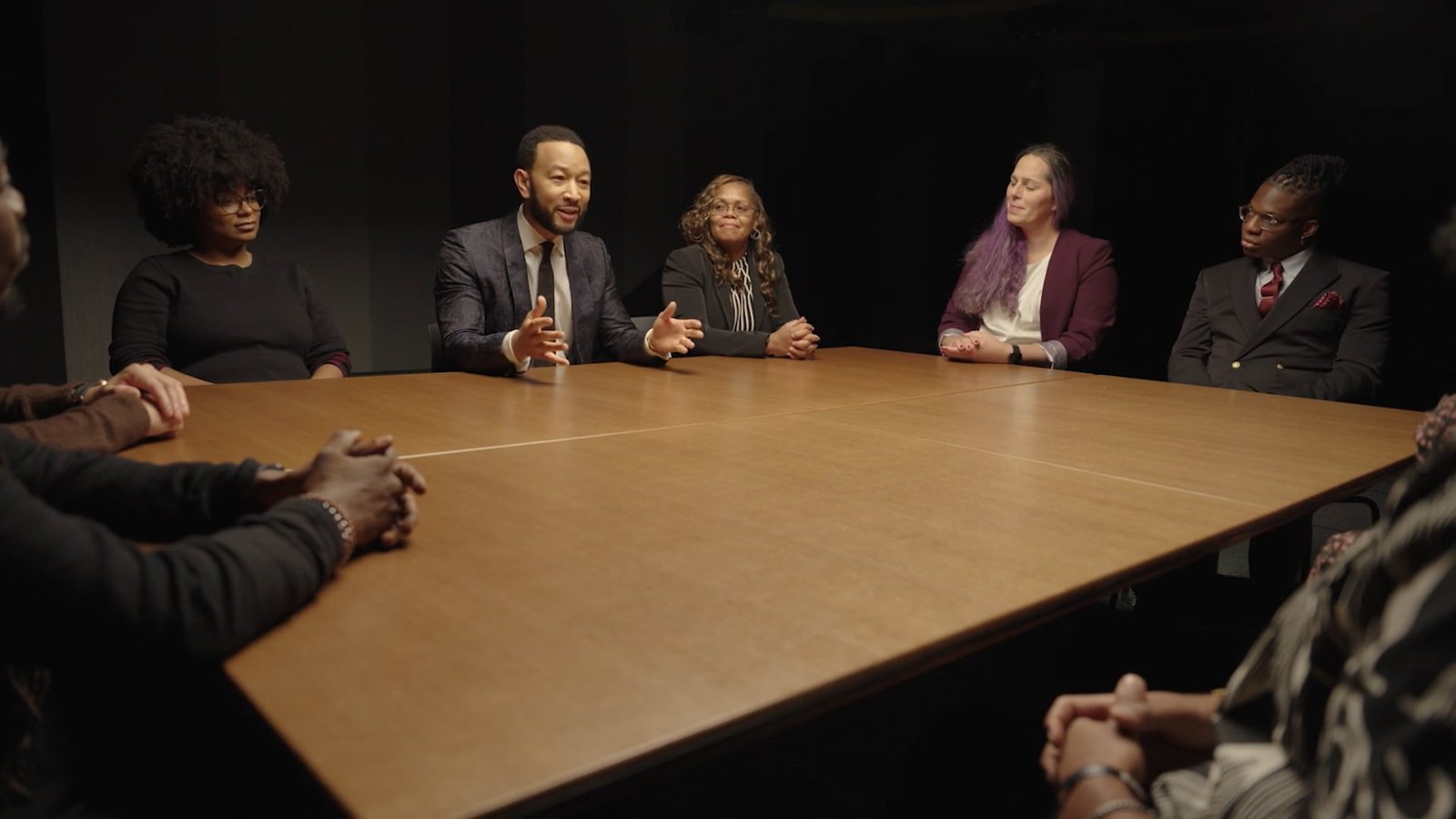 Jessica Hicklin seated at a long wooden table in a dark room with John Legend and other Unlocked Futures social entrepreneurs.