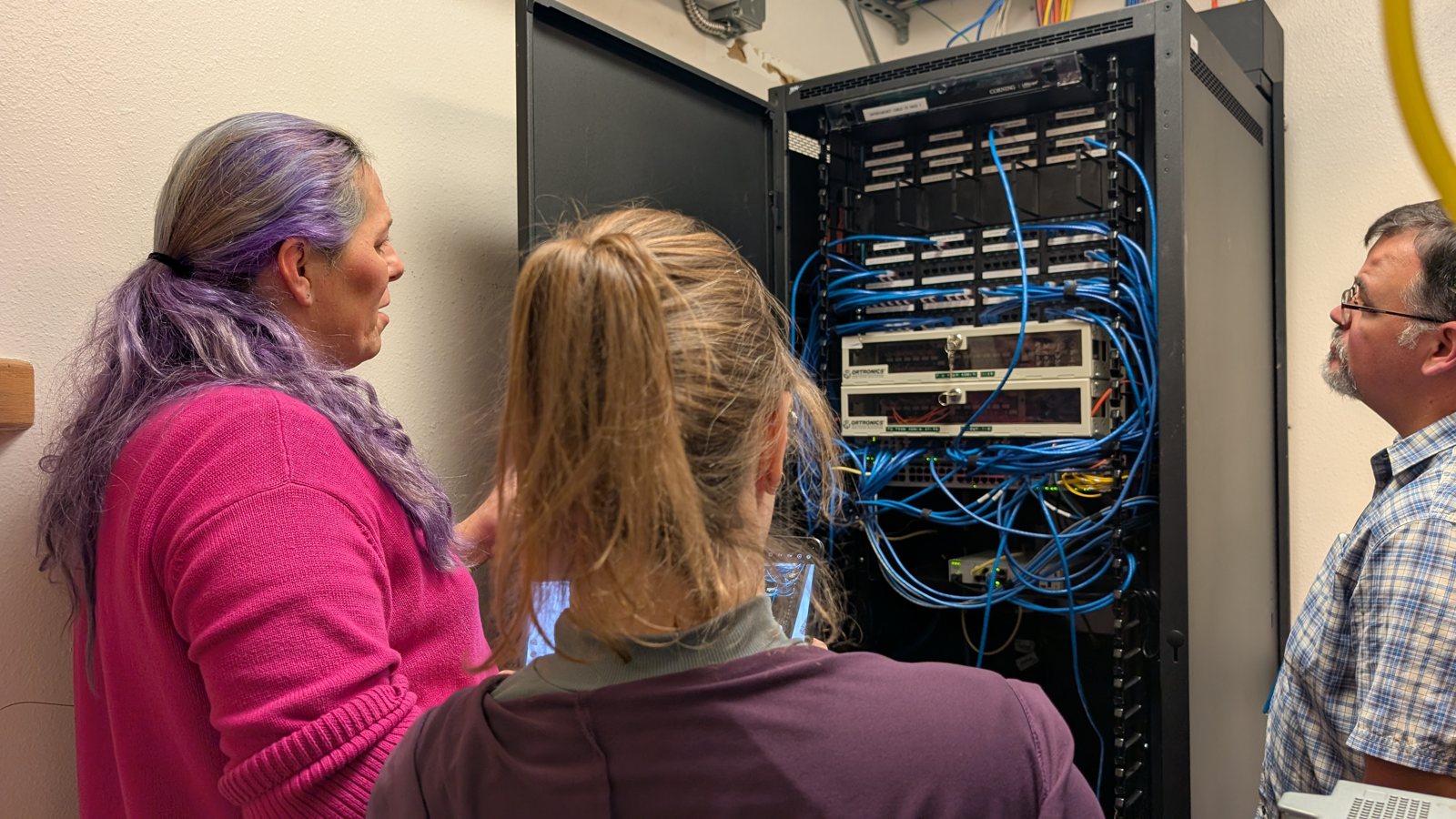 Jessica Hicklin and two colleagues looking into a network rack of blue cables.
