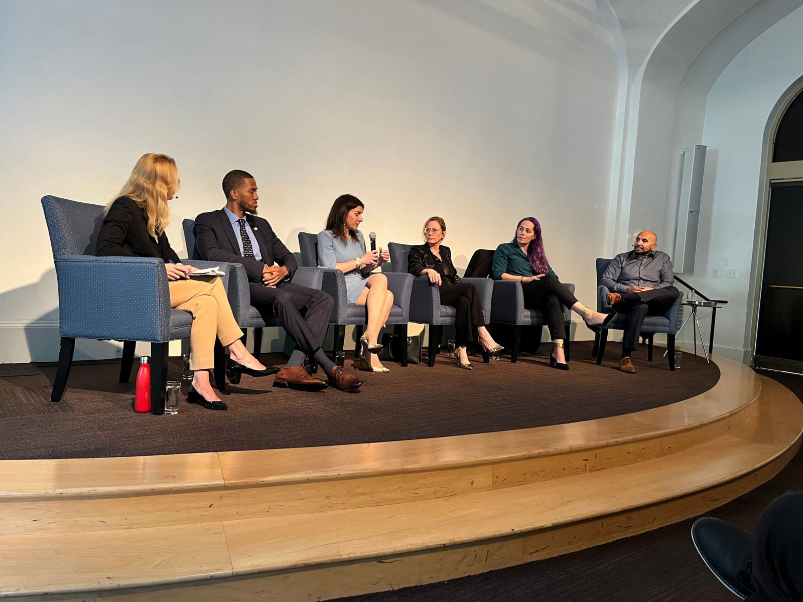 Six people seated on a stage during a panel discussion, Jessica Hicklin fifth from the left.