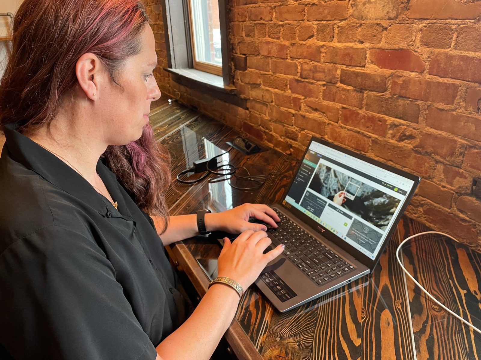 Jessica Hicklin typing on a laptop at a window desk beside a brick wall.